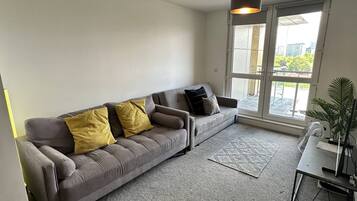 A modern living room with a grey sofa, yellow and black cushions, a grey carpet, a small table with a laptop, and a large window with a view of buildings and trees.