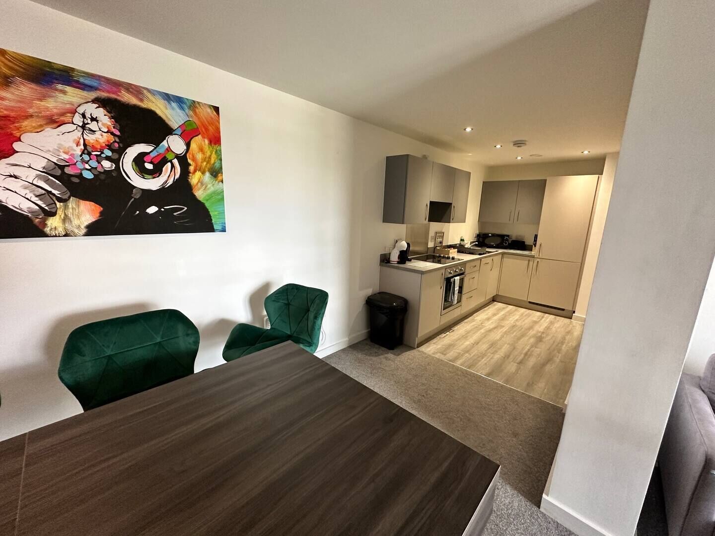 Una cocina moderna con una isla central, un comedor con mesa de madera y sillas verdes, y un colorido cuadro abstracto en la pared.