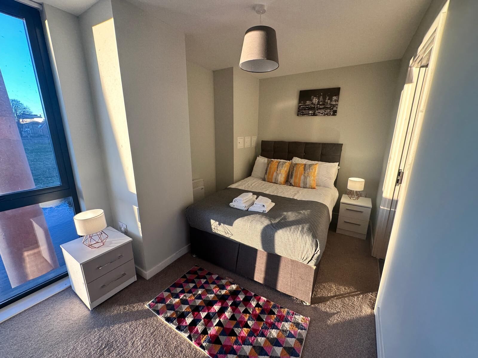 A modern bedroom with a large bed, a bedside table with a lamp, a window with a view of buildings, and a geometric rug.