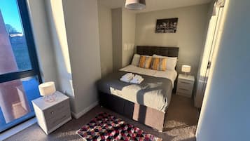 A modern bedroom with a large bed, a bedside table with a lamp, a window with a view of buildings, and a geometric rug.