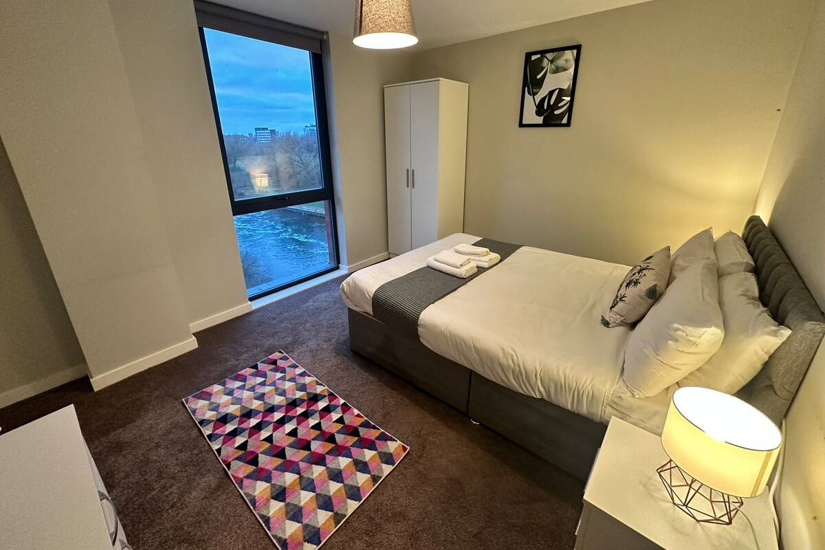 Une chambre d’hôtel avec un grand lit, une armoire, une table de chevet avec une lampe, un tapis coloré et une vue sur la ville par une grande fenêtre.