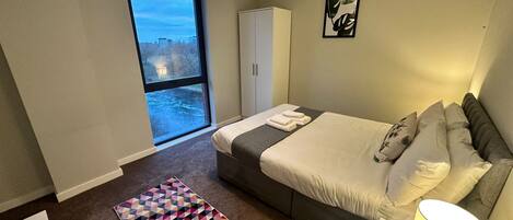A hotel room with a large bed, a wardrobe, a bedside table with a lamp, a colorful rug, and a view of the city through a large window.