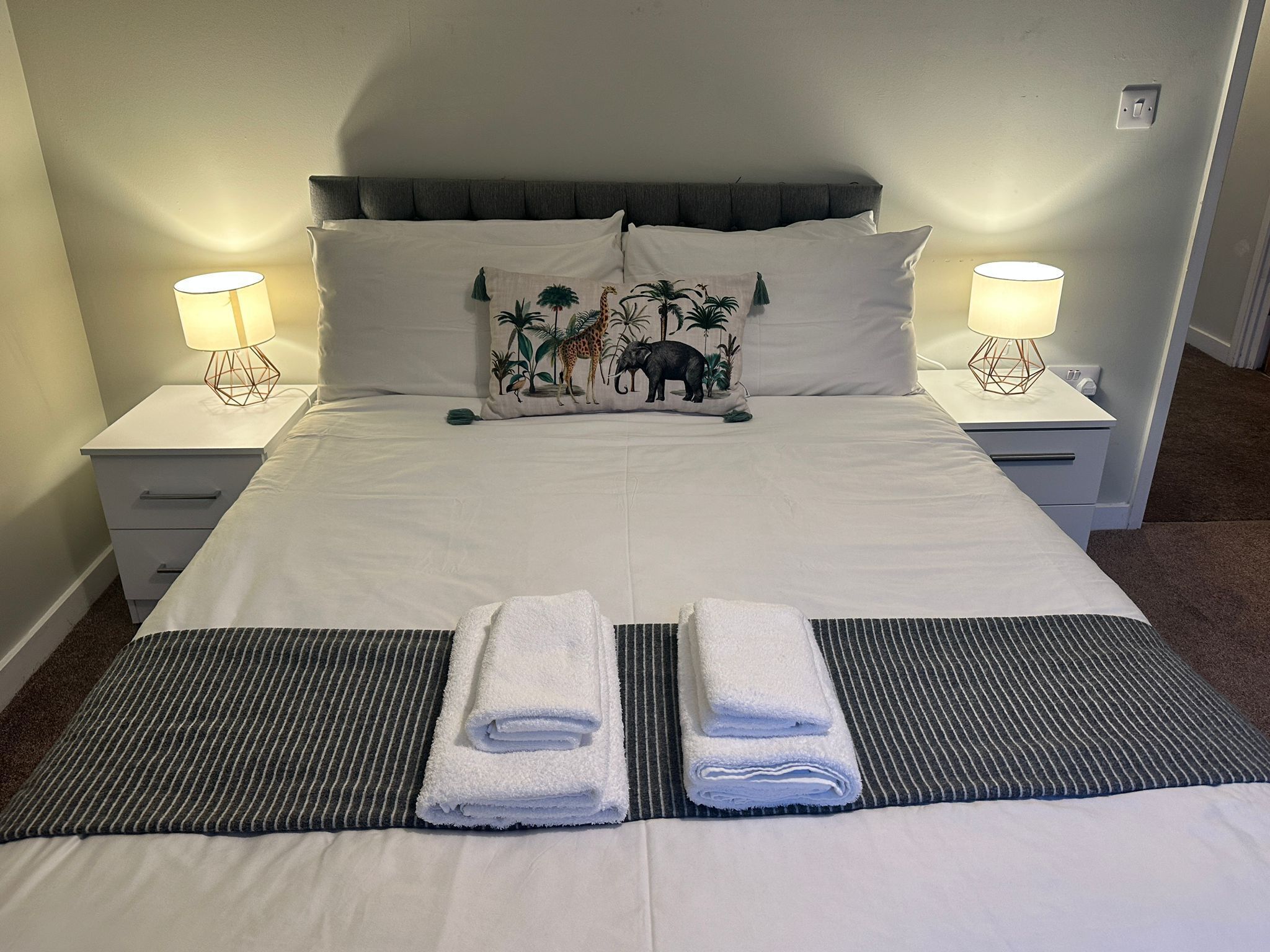 A neatly made bed with white linens, two matching pillows with a tropical print, and two bedside lamps on white nightstands.