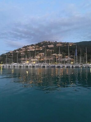 Una marina con varias embarcaciones amarradas, una colina con edificios al fondo y agua tranquila que refleja las barcas.