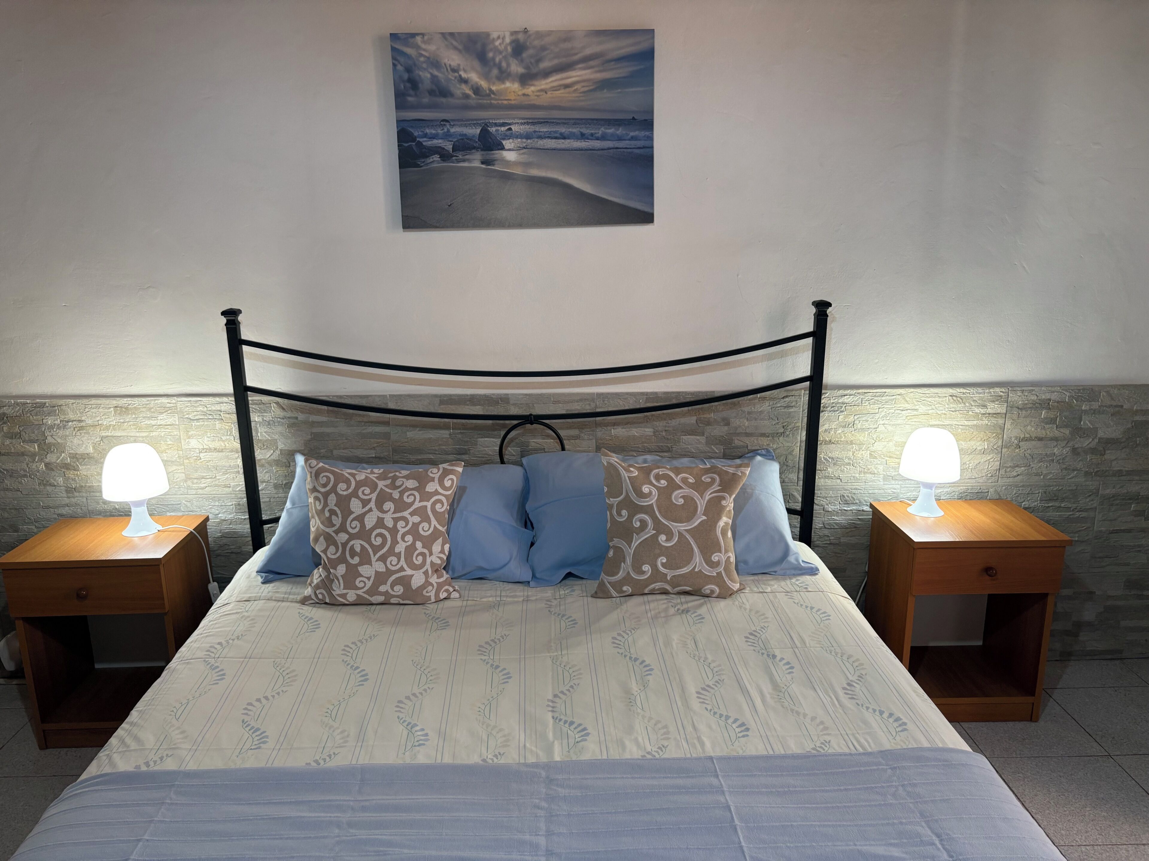 Ein Bett mit Kopfteil, zwei Nachttische mit Lampen, gemusterte Kissen und ein Bild an der Wand.