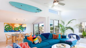 A bright living room with a teal sofa, colorful cushions, a surfboard on the wall, and a large window with a view.