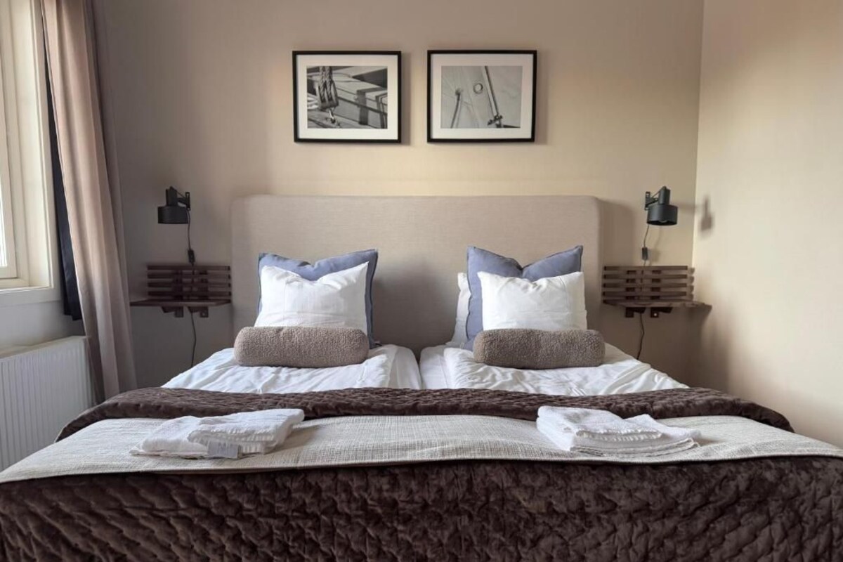 Een tweepersoonsbed met een bruine gewatteerde spreideken, twee nachtkastjes met lampen en twee ingelijste foto's aan de muur.