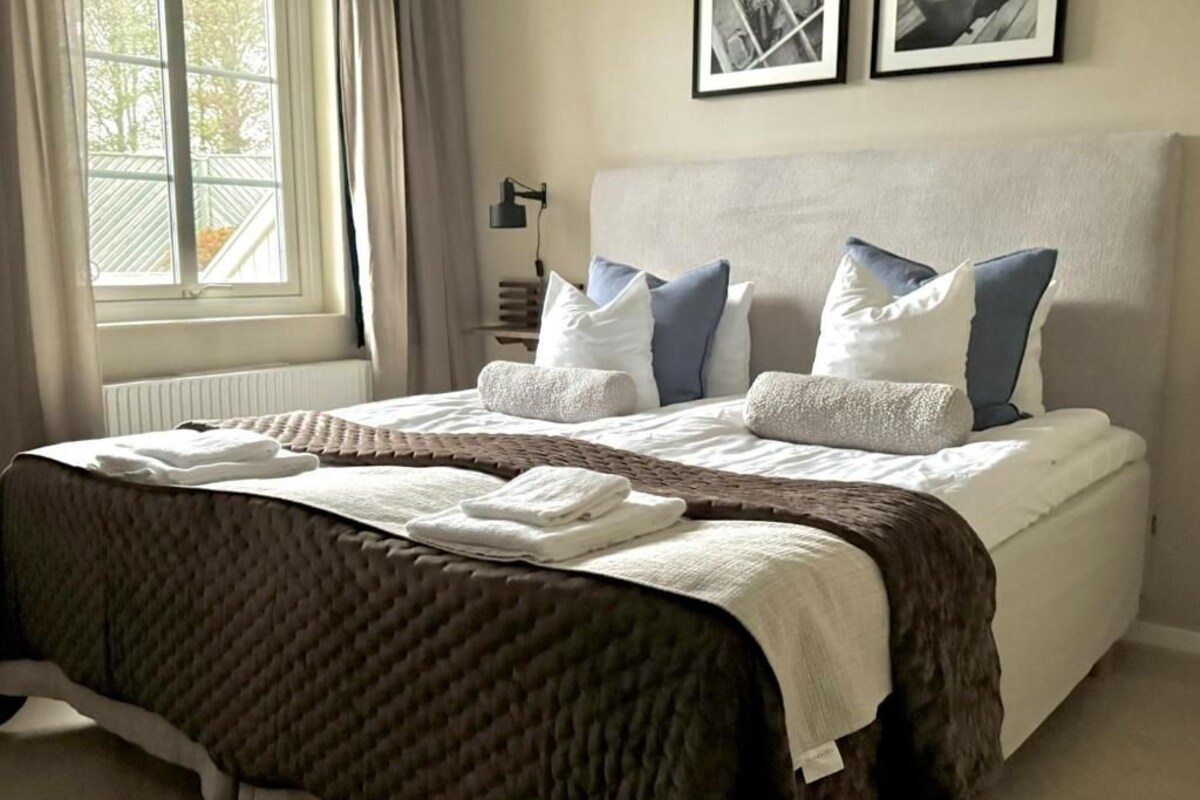 Een net opgemaakt bed met een bruine quilt, witte kussens en een witte deken. Boven het bed hangen twee ingelijste foto's aan de muur.