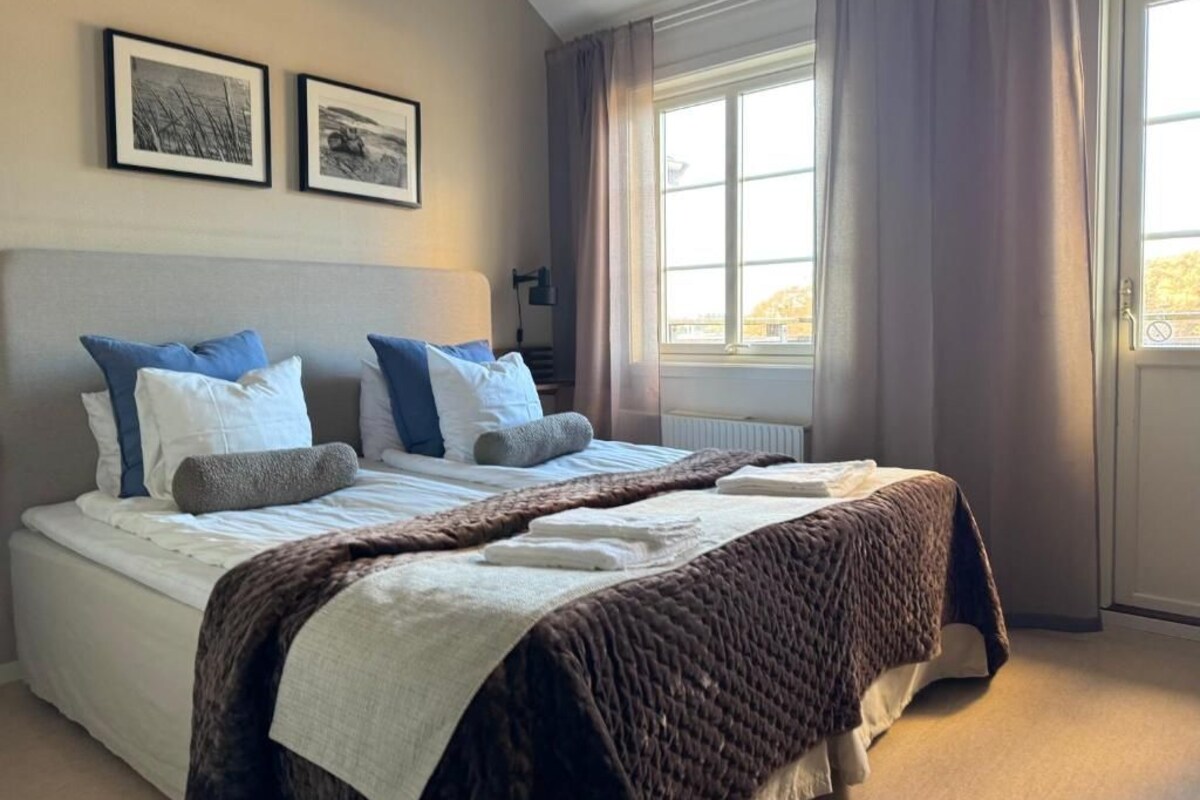 Een net opgemaakt bed met een bruin sprei, witte lakens en blauwe kussens. Boven het bed hangen twee ingelijste foto’s.