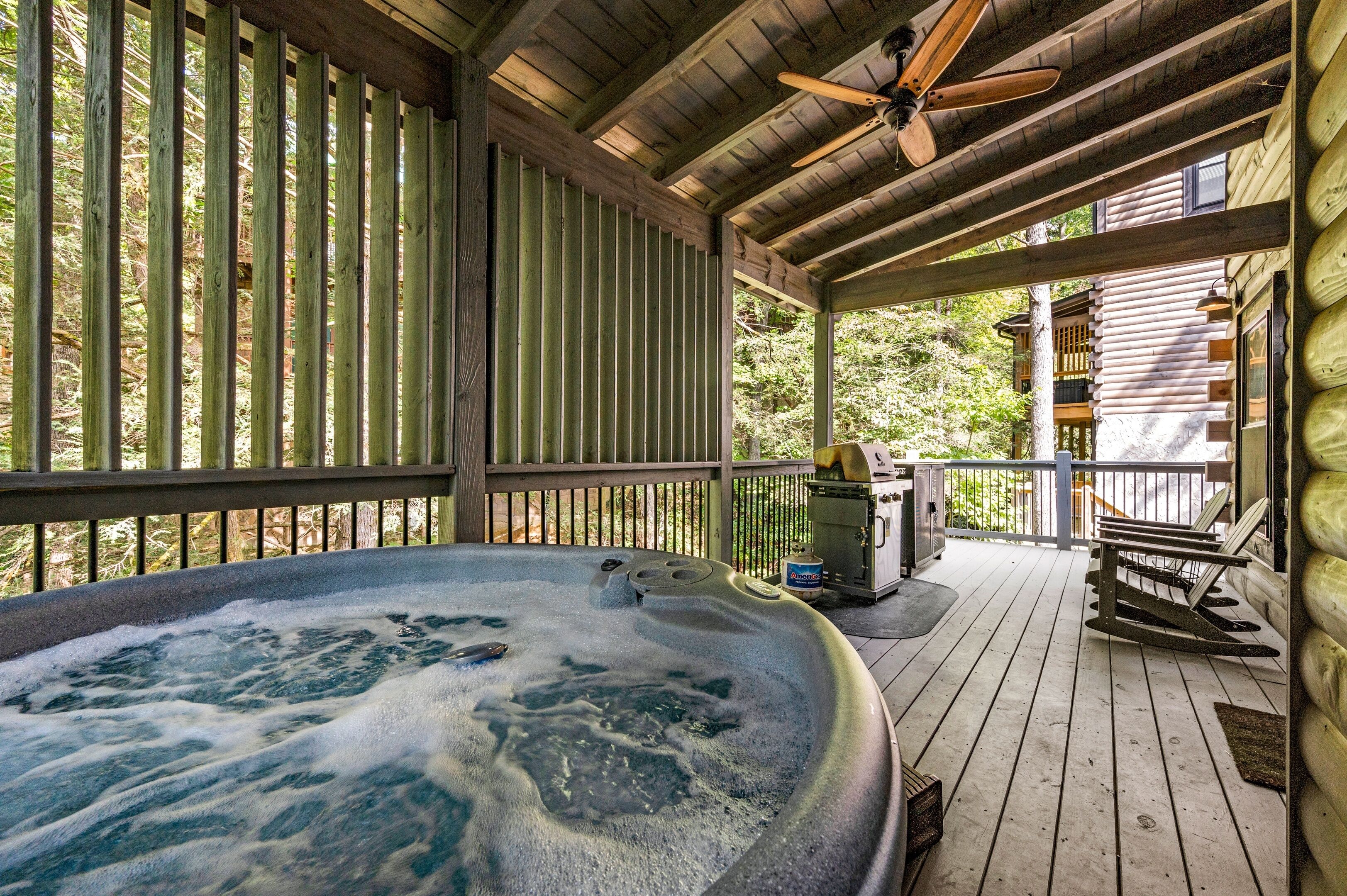 A hot tub on a wooden deck with a ceiling fan, surrounded by wooden railings and a view of a cabin.