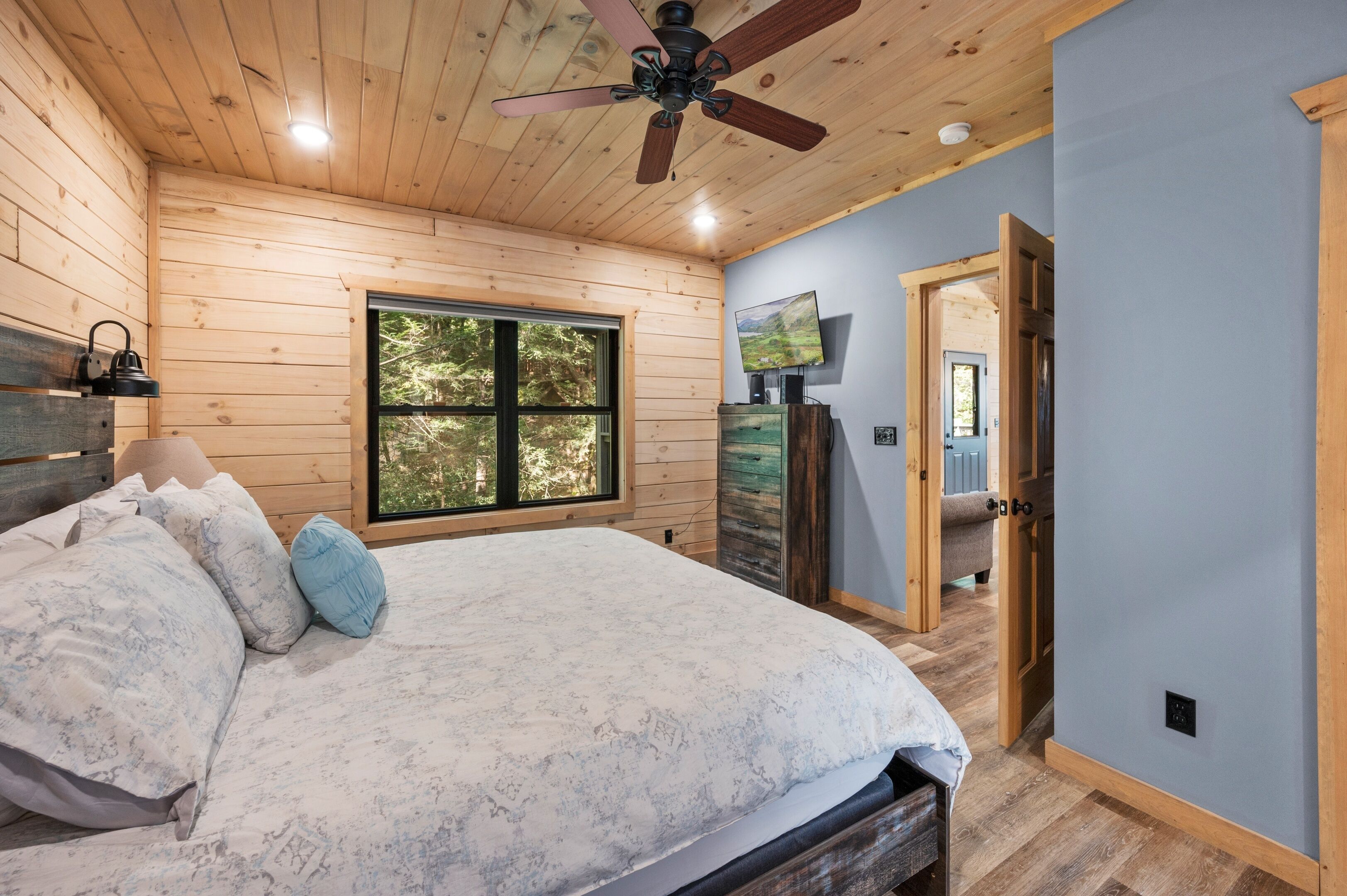 A bedroom with a wooden ceiling, a bed with a white and blue patterned bedspread, a ceiling fan, and a window with a view of trees.