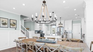 A dining area with a large wooden table and chairs, a chandelier, and a kitchen in the background. - Fish-n-chips By The Sea 6 Bedroom Home by RedAwning (Supply)