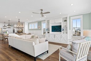 Un salón amplio con un sofá blanco grande, una silla de madera y un televisor sobre un mueble blanco.