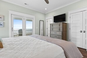 A bedroom with a large bed, a dresser, a TV, and a view of the beach through a door.