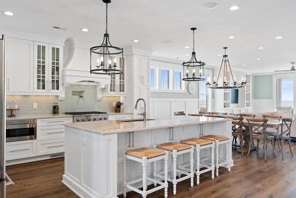 Una cocina amplia con muebles blancos, una isla central con taburetes, iluminación colgante y vistas al área de comedor.