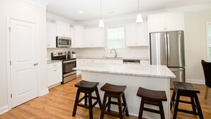 A modern kitchen with white cabinets, stainless steel appliances, and a granite countertop. There are three wooden stools at the island. - Great Location! 4 Bed / 4.5 Bath House in Pinehurst! (Pinehurst)