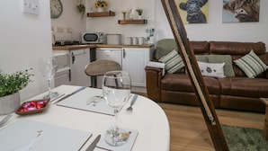 A modern kitchen with a microwave, sink, and wooden countertop. There is a dining area with a table set for two, a wine glass, and a bowl of chocolates. A brown leather sofa is in the living area with decorative pillows. - High Wreah Bolt Hole (Whitehaven)