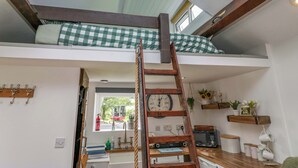 A loft bedroom with a wooden ladder, a bunk bed, a kitchen area with a microwave, and a window with a view of a red vehicle outside. - High Wreah Bolt Hole (Whitehaven)