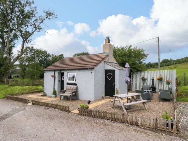 A small, single-story building with a sloped roof, a wooden bench, a picnic table, and a heart-shaped decoration on the door. - High Wreah Bolt Hole (Whitehaven)