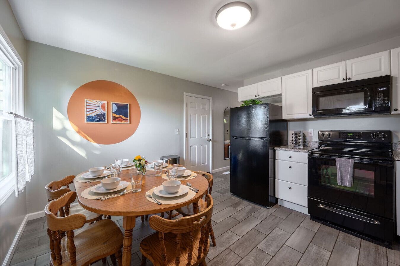 A modern kitchen with white cabinets, a black refrigerator, a microwave, and an oven. There is a dining table with chairs and a vase of flowers.