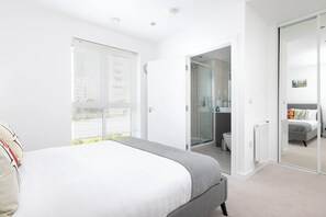 A bedroom with a bed, a window with blinds, a bathroom with a shower, and a closet with a mirror. - Skyvillion - Olympic Flat-excel o2 Arena Westfield (London)