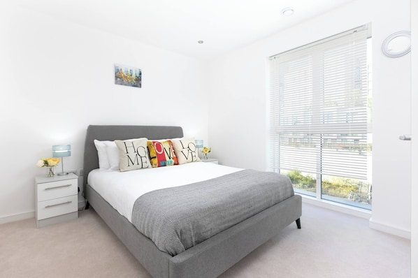 A bedroom with a bed, bedside tables, a window with blinds, and a small painting on the wall.