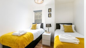 Hotelkamer met twee bedden, elk met een gele deken en witte handdoeken. Er is een nachtkastje met een lamp en een kleine plant.