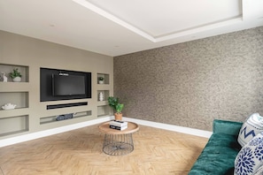 A modern living room with a flat-screen TV, a round coffee table with books, a green sofa, and a patterned wall.