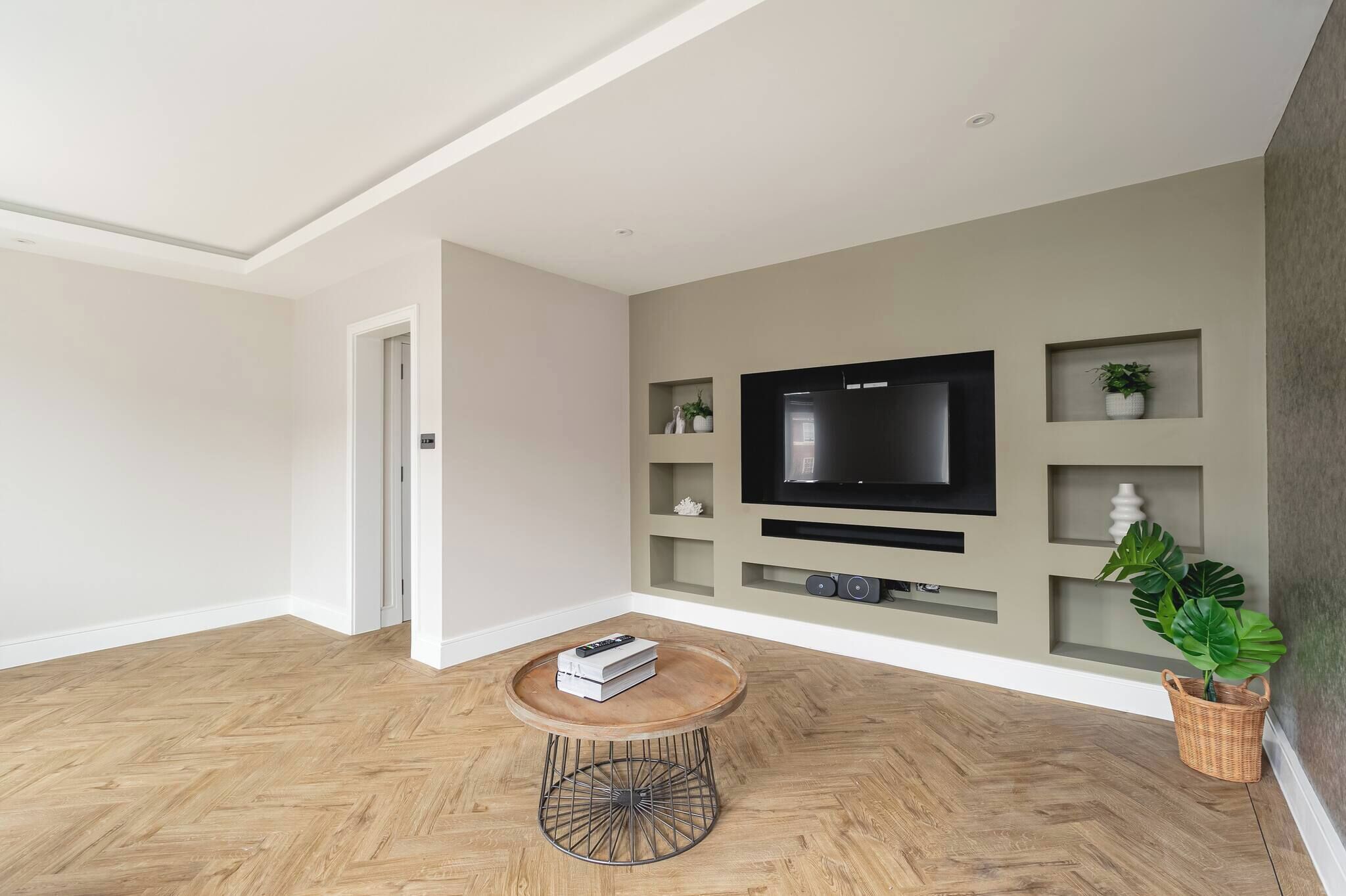 A modern living room with a wooden herringbone floor, a round coffee table with a metal base, a flat-screen TV mounted on the wall, and a potted plant.