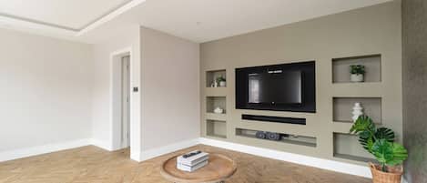 A modern living room with a wooden herringbone floor, a round coffee table with a metal base, a flat-screen TV mounted on the wall, and a potted plant.