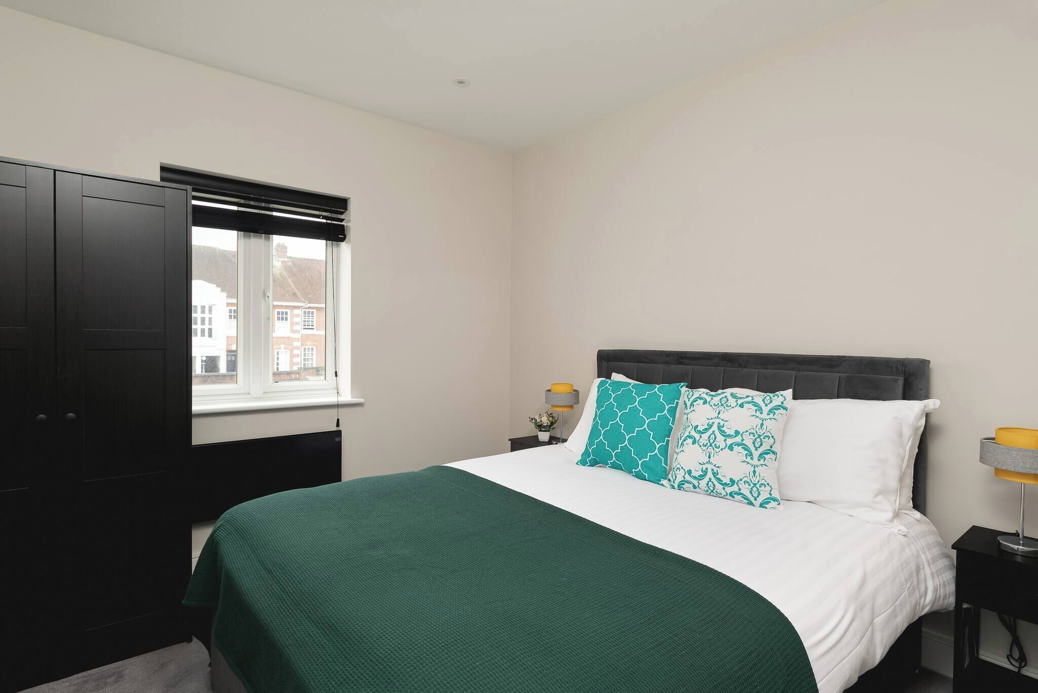 A bedroom with a bed, a window, a wardrobe, and a nightstand with a lamp.