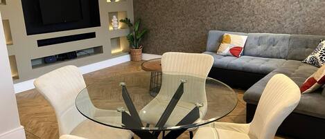 A modern living room with a glass-top coffee table, four white cushioned chairs, a grey sofa with decorative pillows, a flat-screen TV mounted on the wall, and a small potted plant on a shelf.