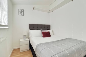 A bedroom with a bed, a bedside table, a nightstand, a wall-mounted shelf, and a window with blinds. - Skyvillion - Valley Way - 3bed House In Stevenage (Stevenage)
