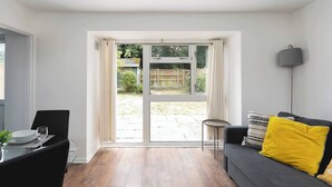 A room with a dining table, chairs, a sofa with cushions, a lamp, and a sliding glass door leading to an outdoor area. - Skyvillion - Lush 2 Bed Hydean Way House & Garden (Stevenage)