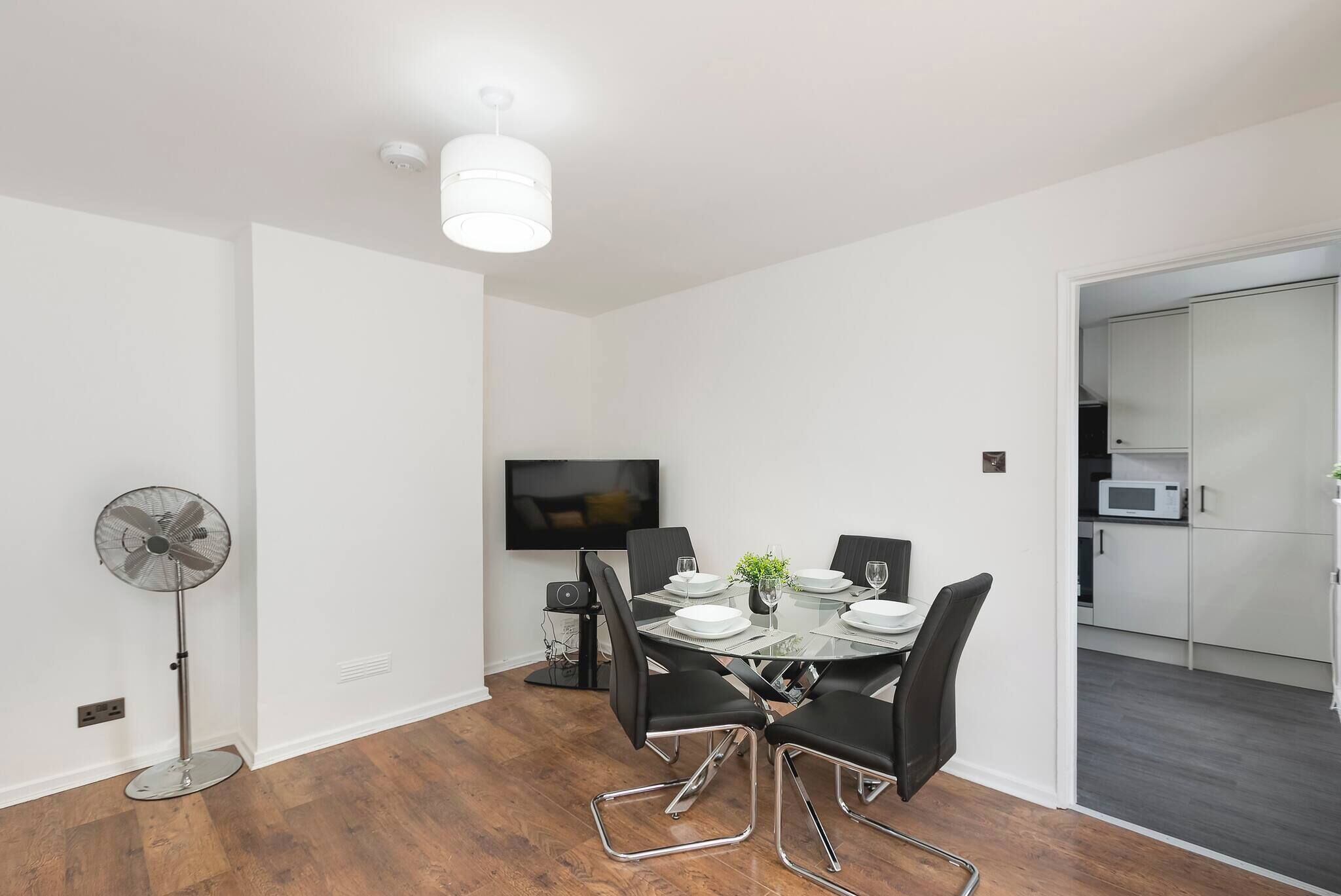 A modern dining area with a table set for four, a standing fan, a flat-screen TV, and a kitchen area visible through an open door.