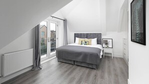 A modern bedroom with a sloped ceiling, a large bed, a nightstand, and a view through a glass door. - Skyvillion -vineyard Court- Enfield 1bed Wbalcony (Enfield)