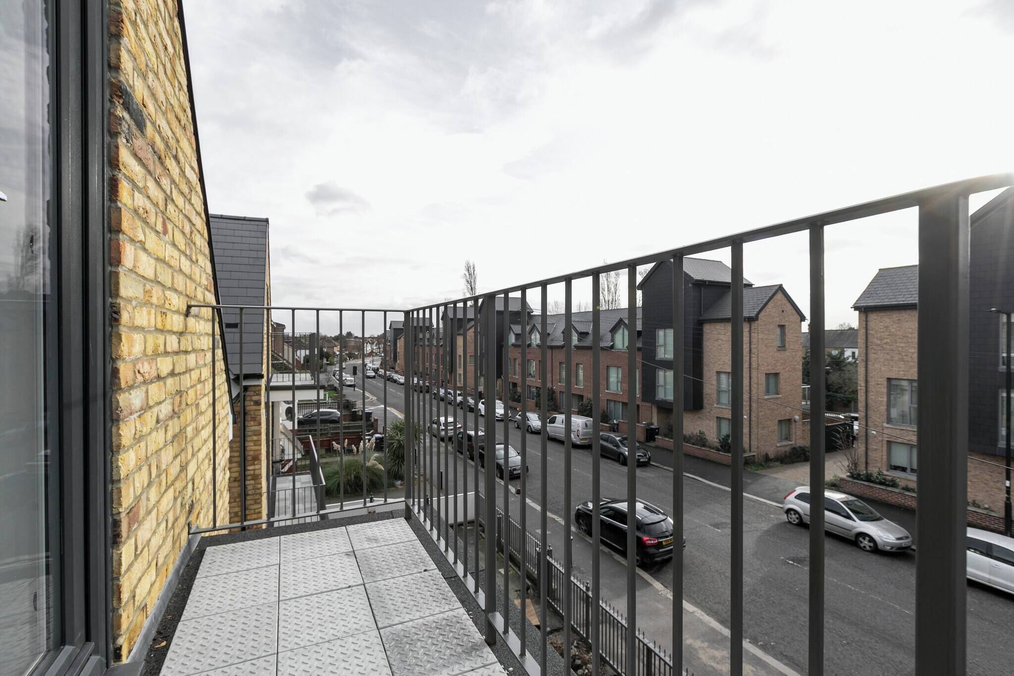 A balcony with a view of a residential street, a brick building, and parked cars.