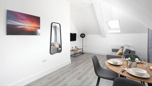 A modern dining area with a wooden table set for two, a grey sofa, and a large painting of a sunset. - Skyvillion -vineyard Court- Enfield 1bed Wbalcony (Enfield)