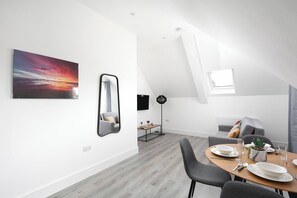 A modern dining area with a wooden table set for two, a grey sofa, and a large painting of a sunset.