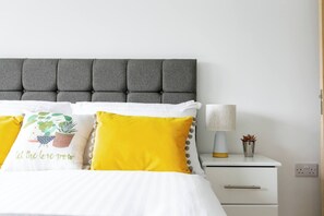 Una cama bien hecha con almohadas blancas y amarillas, un cabecero gris con botonadura y una lámpara de noche con una planta pequeña en el mesita de noche.