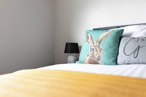 Uma cama com um cobertor amarelo, uma luminária preta e duas almofadas com estampa de coelho e design parcial de texto.