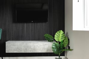 Quarto moderno com televisão de tela plana presa a uma parede escura, um banco cinza e uma grande planta verde.
