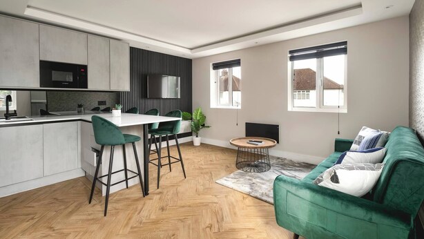 A modern kitchen with a microwave, bar stools, and a dining table. There is a green sofa with patterned cushions and a small round table with a plant. - Skyvillion -1bed Cockfosters Near Alexandra Palace (Barnet)