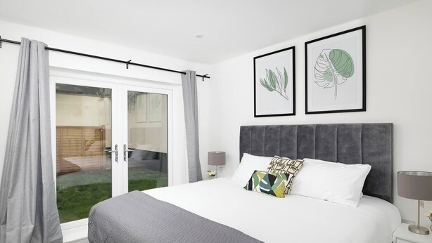 Quarto com cama, mesa de cabeceira, abajur, janela com cortinas e dois quadros com ilustrações de plantas na parede.