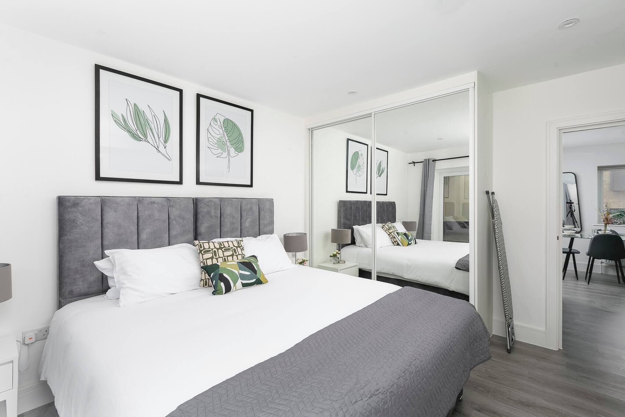A modern bedroom with a large bed, a mirror, and framed botanical prints on the wall.