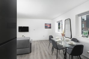 Ett modernt matområde med ett glasbord, grå stolar, en platt-TV och en tavla som föreställer en stadsbild.