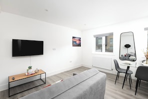 Una sala moderna con televisor de pantalla plana, una mesa de centro de vidrio, un sofá gris, un comedor con mesa y sillas de vidrio, y un espejo grande.