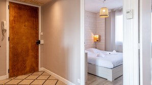Een hotelkamer met een houten deur, een bed met wit beddengoed, een wandlamp en een houten vloer.