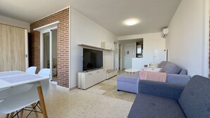 Ein modernes Wohnzimmer mit Flachbildfernseher, grauem Sofa, kleinem Tisch und Essbereich mit Stühlen.