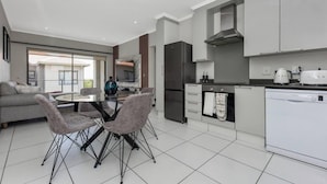 Una cocina moderna con lavavajillas integrado, un refrigerador grande y un microondas. Hay una zona de comedor con una mesa de vidrio y cuatro sillas.
